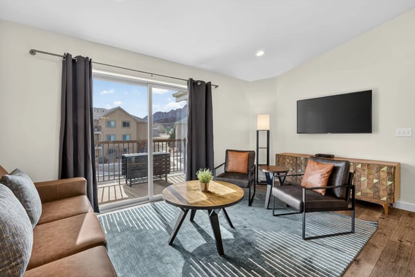 Bright living room with leather seating, round wood table, wall-mounted TV, and glass doors opening to mountain view balcony
